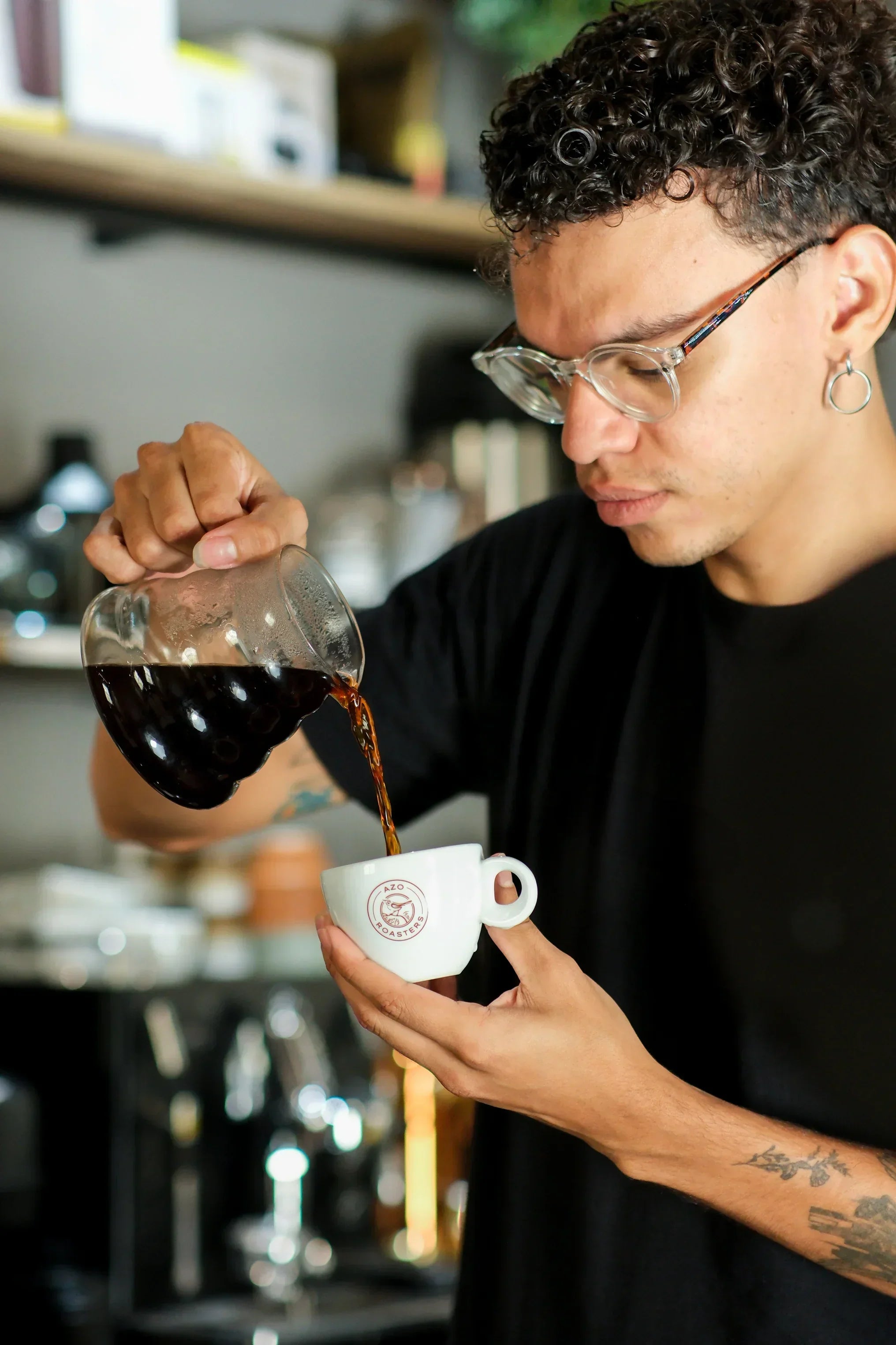 Barista jovem com óculos e tatuagem servindo café em xícara branca da Azo Roasters